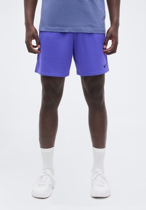 Άνδρας που φοράει μωβ αθλητικά σορτς, άσπρες κάλτσες, άσπρα αθλητικά παπούτσια, στέκεται όρθιος μπροστά σε ένα απλό φωτεινό φόντο.