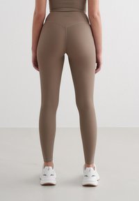 Leggings marron côtelés à taille haute et tissu texturé, associés à des chaussures de sport blanches sur un sol clair.