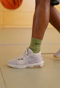 Bijele košarkaške tenisice s mrežastim detaljima, sivi akcenti i vidljiva zračna amortizacija, uparene sa zelenim rebrastim čarapama, na terenu za gimnastiku.
