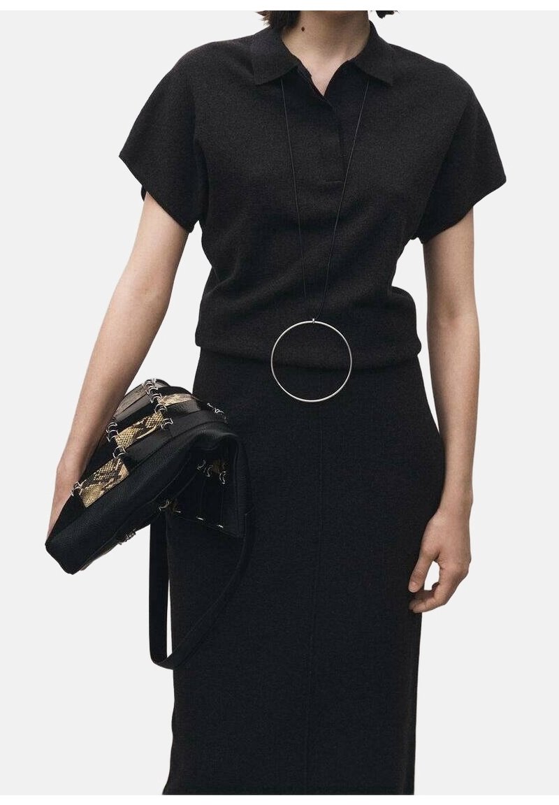 Femme portant une robe noire à col avec manches courtes, un grand pendentif circulaire autour du cou, tenant un sac à main noir à motif de peau de serpent.