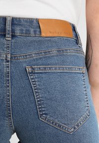 Niebieskie dżinsy o wysokim stanie, widoczne przeszycia, skórzana metka z tyłu z napisem "Noisy May" oraz detale na tylnej kieszeni.