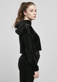 Svart cropped hettegenser laget av myk fløyelsmateriale. Har hette, lange ermer og elastiske mansjetter, med en glatt tekstur og figursydd design.