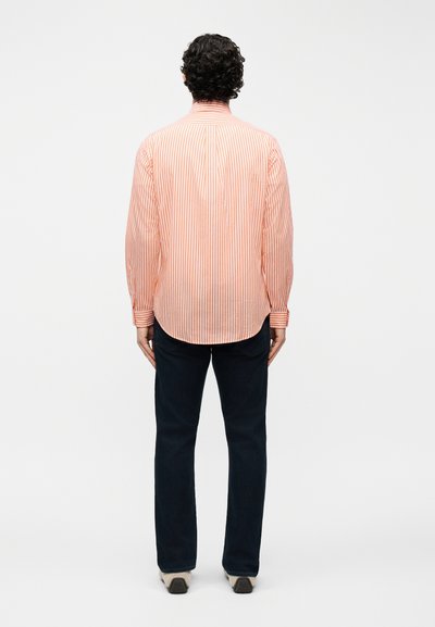 Άνδρας με σγουρά σκούρα μαλλιά, φορώντας πορτοκαλί και άσπρη ριγέ μακρυμάνικη μπλούζα, σκούρο παντελόνι και άσπρα παπούτσια, στέκεται γυρισμένος με την πλάτη.