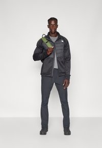 Chaqueta aislante negra con cierre frontal, camiseta gris, pantalones oscuros y botas negras. Lleva un bolso verde al hombro. No se ven patrones.