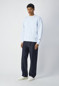 Sudadera azul clara con puños y dobladillo acanalados, que presenta un pequeño logo en el pecho. Combinada con pantalones de mezclilla oscuros y zapatillas blancas.