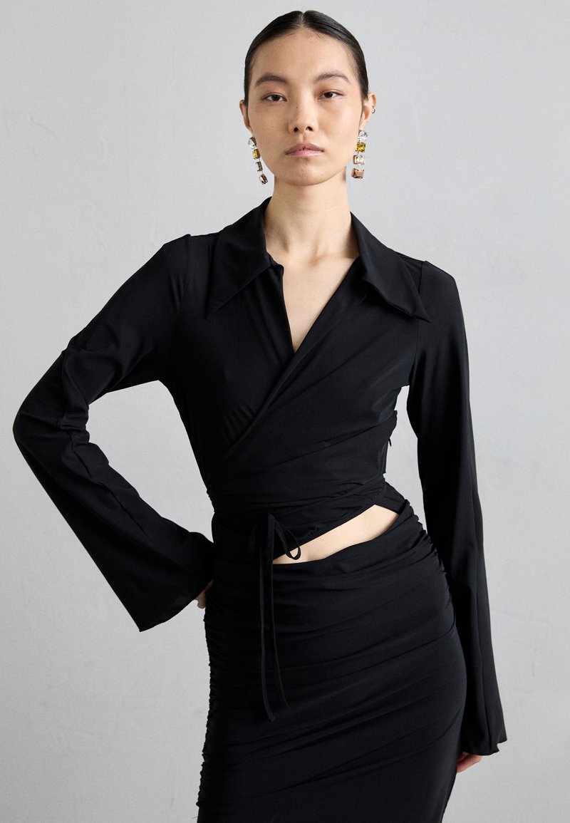 Zwarte wikkeltop met lange, uitlopende mouwen, voorzien van een diepe kraag en een gestrikt middel. Glad, rekbaar materiaal benadrukt het aansluitende ontwerp.