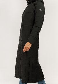 Czarny długi pikowany płaszcz z przednim zamkiem błyskawicznym, kapturem i naszywką z logo na górnej części rękawa, noszony na niebieskich dżinsach.