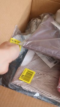 Scatola di cartone piena di vari oggetti, tra cui un tessuto a righe, imballaggi in plastica e codici a barre visibili su etichette.