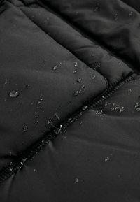 Des gouttes d'eau perlent sur un tissu matelassé noir près d'une fermeture éclair, indiquant un matériau résistant à l'eau ou imperméable.
