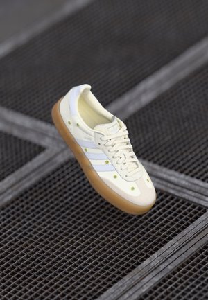 Hvid Adidas Samba sneaker med brun sål og små grønne blomstermønstre, der svæver over et metalgittergulv.