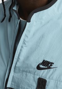 Ljusblå Nike-jacka med svart dragkedja, svarta kanter och broderad logotyp. Materialet verkar lätt och har en slät textur.