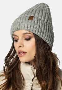 Berretto lavorato a maglia grigio con texture a coste e un piccolo tag in pelle marrone; indossato con una giacca a vento chiara.