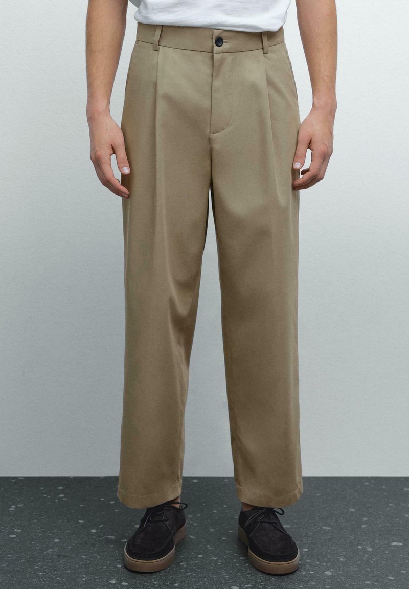 Persoon in crème kleuren, op maat gemaakte broek met een wit shirt en donkere schoenen, staand op een gestructureerd donkere ondergrond tegen een effen achtergrond.