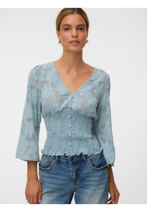 Femme vêtue d'un chemisier bleu clair à texture florale avec des boutons et une taille smockée, associé à un jean denim bleu sur un fond blanc uni.