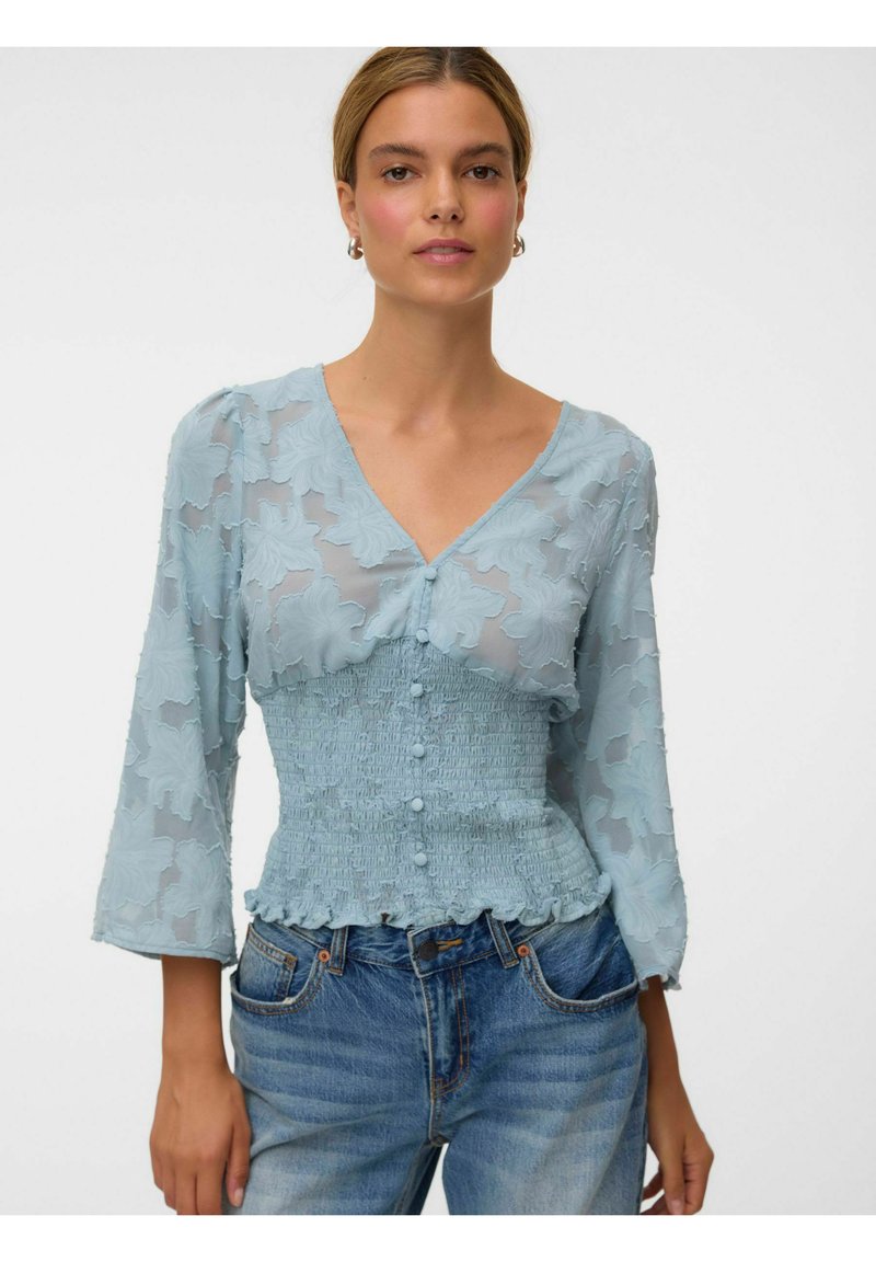 Femme vêtue d'un chemisier bleu clair à texture florale avec des boutons et une taille smockée, associé à un jean denim bleu sur un fond blanc uni.