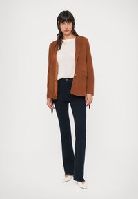 Blazer de veludo cotelê castanho, camisola creme, jeans escuros com corte flare e sapatos brancos pontiagudos. O blazer apresenta um único botão e bolsos frontais.