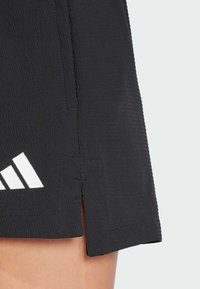 Black athletic skirt with textured fabric, featuring a side slit and white logo accent. Visible stitching details enhance the design.