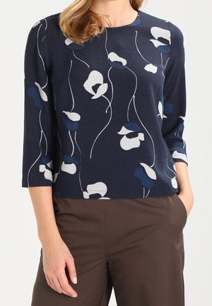 Femme portant un haut bleu marine à manches longues avec des motifs floraux blancs et bleu foncé et un pantalon marron, debout avec les mains partiellement dans les poches.
