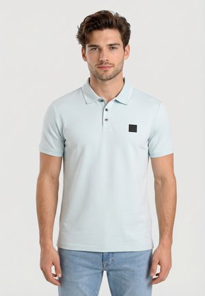 Uomo con capelli castani che indossa una polo azzurra a maniche corte con tre bottoni e jeans blu, in piedi davanti a uno sfondo chiaro uniforme.