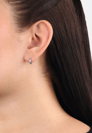 Nahaufnahme eines Ohres einer Person mit glattem, dunkelbraunem Haar, die einen kleinen silbernen Ringohrring am Ohrläppchen trägt.