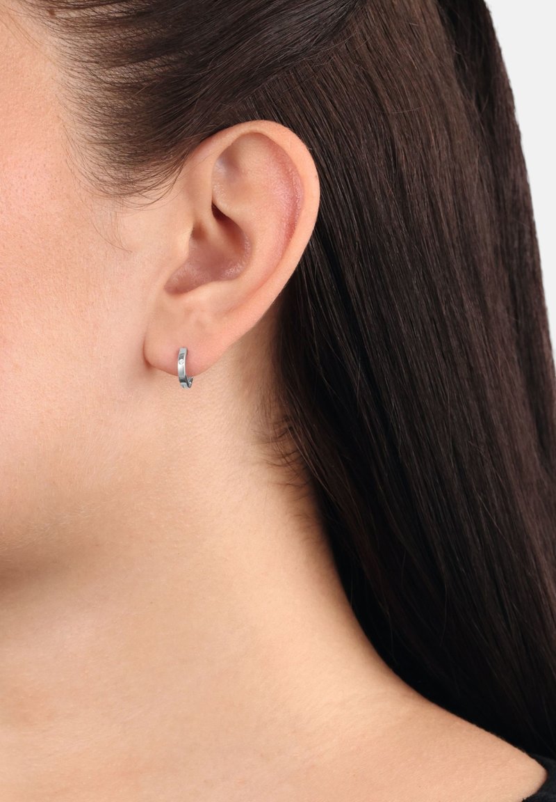 Primer plano de la oreja de una persona con cabello lacio castaño oscuro, que lleva un pequeño pendiente en forma de aro de plata en el lóbulo de la oreja.