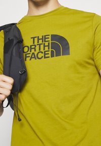 Groene T-shirt met een groot zwart logo van "THE NORTH FACE." De stof lijkt zacht; het model houdt een zwart accessoire vast met een trekkoord.