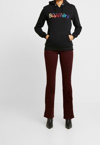 Černá mikina s kapucí s barevným nápisem „Superdry“. Kombinováno s vínovými rozšířenými kalhotami a černými kotníkovými botami. Jednoduchý, ležérní outfit.