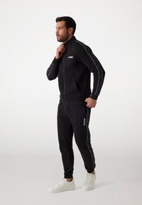 Schwarzer Trainingsanzug mit einer Reißverschlussjacke, die über Fronttaschen und weiße Details verfügt, kombiniert mit passenden schmalen Hosen und weißen Sneakers.