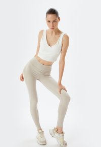 Lichtbeige leggings met een hoge tailleband, gecombineerd met een witte mouwloze crop top. Het model draagt crèmekleurige en grijze sportschoenen.