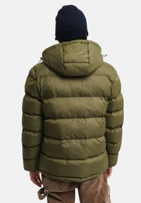 Giubbotto imbottito verde oliva, con cappuccio, design trapuntato e polsini elasticizzati. Indossato con pantaloni cargo marroni, visto da dietro.