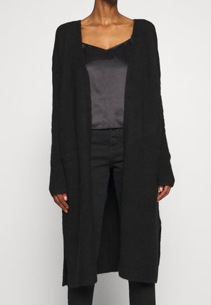 Femme portant un long cardigan noir ouvert sur un caraco noir en satin et un pantalon taille haute noir, debout devant un fond uni.
