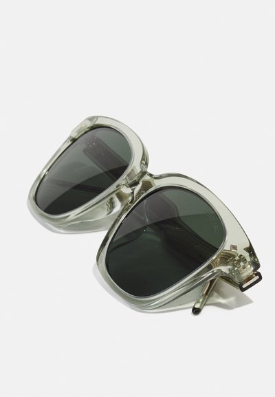 Ochelari de soare mari din plastic transparent, cu lentile verzi închise și accente argintii. Formă unică, unghiulară, cu un finisaj lucios.