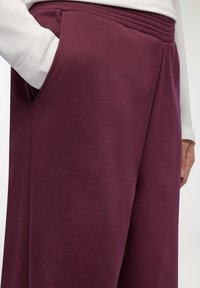 Burgundowe dresy z miękkiej tkaniny, szeroką nogawką, elastycznym paskiem w talii i detalem w postaci kieszeni po bokach. Materiał wygląda na komfortowy i casualowy.