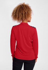 Rotes Sportoberteil mit langen Ärmeln und hohem Kragen, mit schwarzen gemusterten Akzenten an den Schultern und einer glatten Stofftextur.