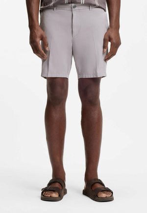 Mężczyzna w jasnoszortkich spodenkach i ciemnobrązowych sandałach stoi na tle białego tła.