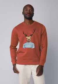 Sweatshirt couleur rouille avec un graphisme d'un cerf portant une chemise rayée bleu et blanc. Texture lisse, design à col rond et poignets côtelés.