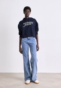 Cappuccio accorciato navy con logo "tommy jeans". Jeans svasati blu chiaro con un lavaggio medio. Sneakers bianche con suola beige. Sfondo neutro.