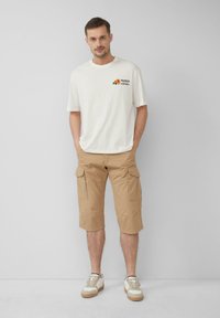 Weißes Baumwoll-T-Shirt mit schwarzem und orangefarbenem Grafikdesign, khakifarbene Cargo-Shorts mit mehreren Taschen und beigem Sneakern mit weißen Akzenten.