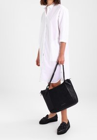 Černá kabelka z broušené kůže se dvěma uchy, zapínáním na zip a texturovanými detaily. Hladká podšívka a minimalistický design. Nošena s bílými šaty.