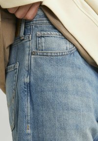 Light blue denim jeans with a faded texture, front pocket detail, and silver metal hardware accents. Partially covered by a beige sweater.