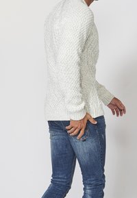 Vit kabelstickad tröja med ribbade ärmslut, bärs över blå jeans. Tröjan har strukturerade mönster och en avslappnad passform, vilket framhäver stickstrukturen.