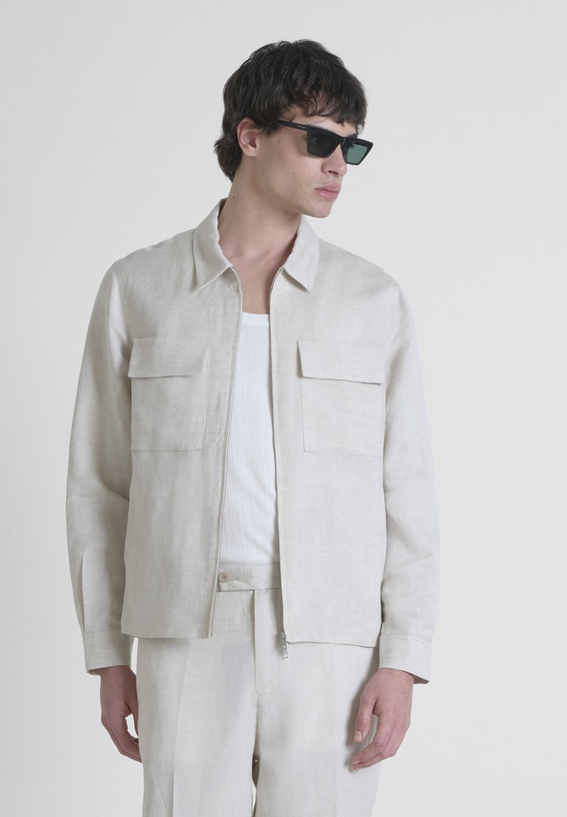 Giacca in lino beige chiaro con due tasche sul petto, indossata su una canotta bianca a coste e pantaloni coordinati. Modella che indossa occhiali da sole scuri.