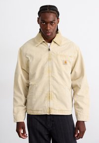 Veste en coton beige avec fermeture éclair, col pointu et poches avant. Présente un petit patch logo sur la poitrine gauche et une coupe décontractée.