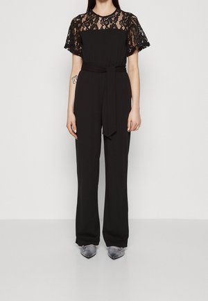 Kvinde iført sort jumpsuit med blonder kortærmer, et bælte i taljen og metalliske spidse sko, stående mod en ensfarvet baggrund.