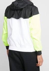 Färgad jacka med svart övre del, vit mittdel och neongröna ärmar. Lättviktsmaterial, med huva och elastiska ärmslut.