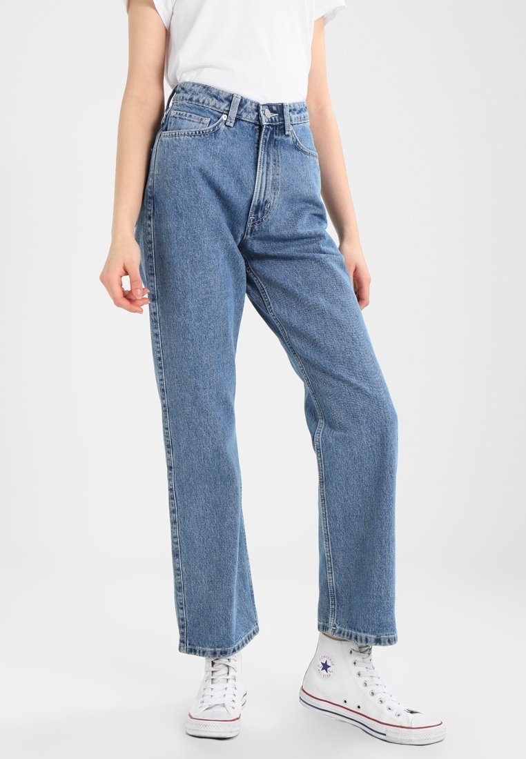 Ljusblå denimjeans med hög midja, avsmalnande ben och subtila blekningar. Kombineras med vita kanvasnikar med en logotyp.