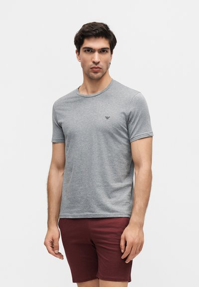 Άνδρας που στέκεται με πρόσωπο μπροστά, φοράει γκρι κοντομάνικη μπλούζα και μπόρντο σορτς σε ένα απλό λευκό φόντο.