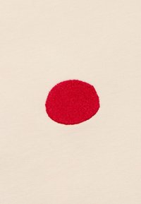 Tessuto beige con un grande cerchio ricamato rosso al centro. La texture è liscia con una finitura morbida.