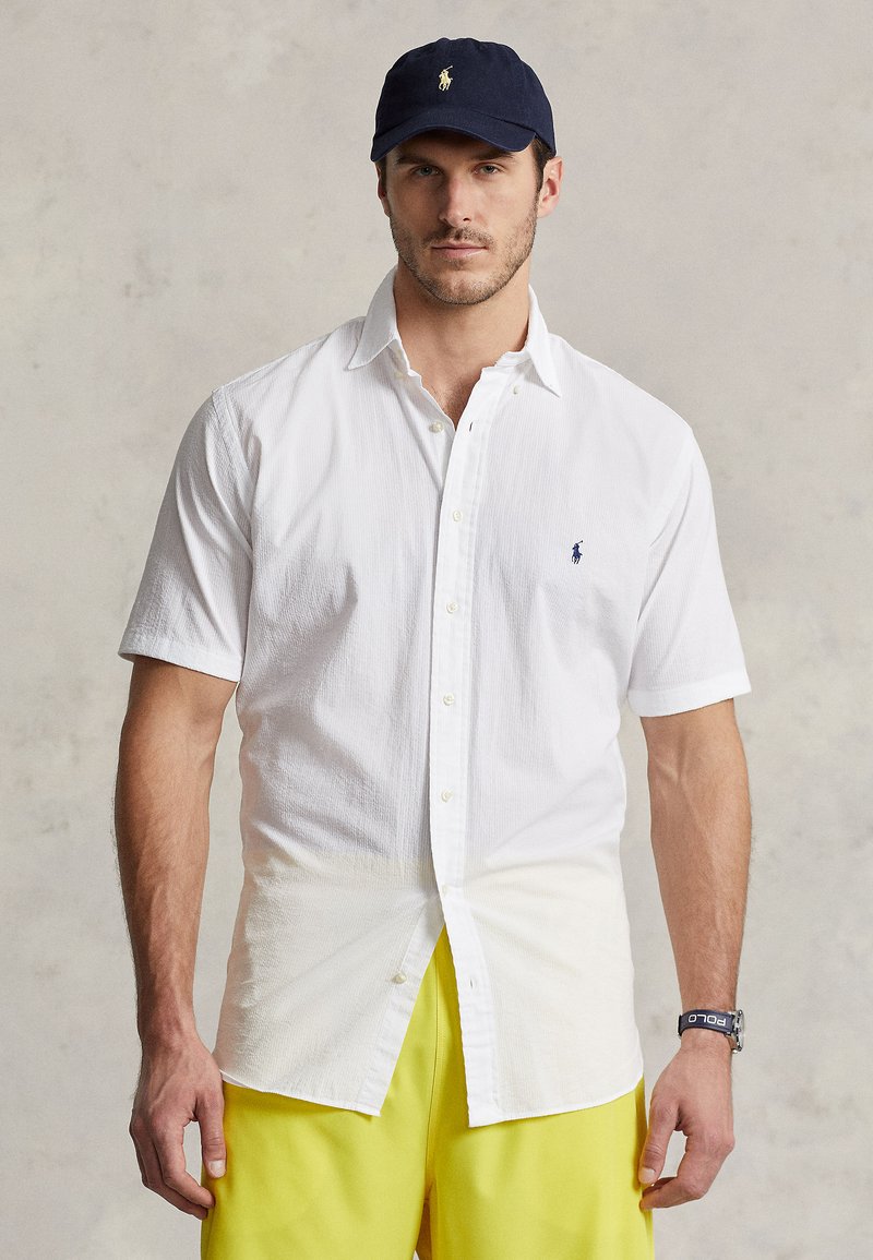 White short-sleeve shirt with a textured finish, featuring a small blue polo logo on the chest, paired with bright yellow shorts and a navy cap.
