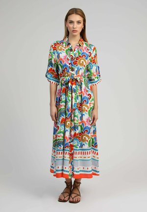 Femme portant une robe midi colorée à motifs floraux, boutonnée, avec une ceinture à la taille et des sandales plates marron à brides sur un fond gris uni.